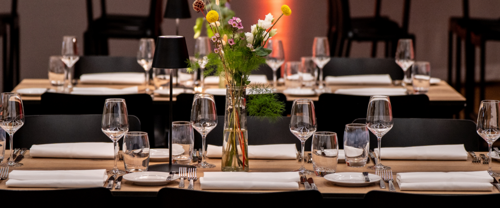 Lange festlich gedeckte Tafel mit Weingläsern, weißen Stoffservietten und Wildblumen-Dekoration – Eventausstattung von Partyladen von Voss