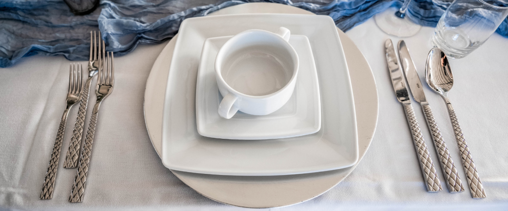 Quadratisches Geschirr-Set mit Tasse und gemustertem Besteck auf blauem Tischläufer – Mietgeschirr von Partyladen von Voss